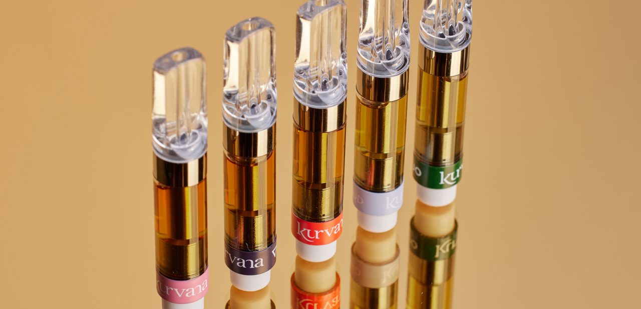 kurvana-originals-1ml-carts | Kurvana | Curated for years. Experienced in moments. Five colorful refillable vape cartridges lined up on a reflective surface with a golden background.